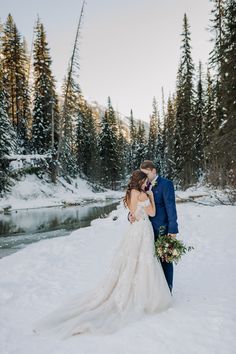 a bride and groom are standing in the snow near a river with pine trees on either side