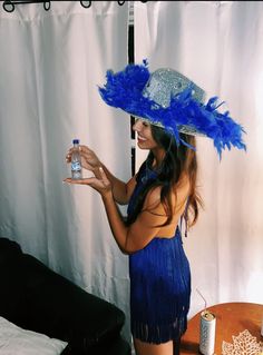 a woman in a blue dress holding a bottle and wearing a hat with feathers on it