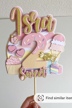 a hand holding up a pink and gold cake topper with the number two on it