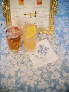 two cocktails are sitting on a table next to a menu and a gold frame