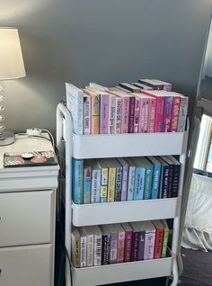 a white book shelf filled with books next to a night stand and lamp on top of a bed