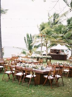 an outdoor wedding reception setup with wooden chairs and tables set up in the grass, surrounded by palm trees