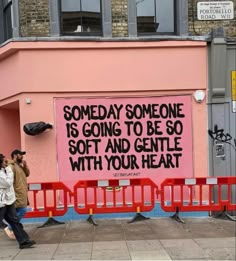 two people walking past a pink wall with writing on it