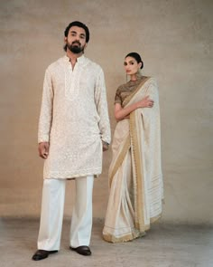 a man and woman standing next to each other in front of a beige wall wearing white clothes