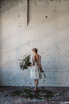 a woman standing in front of a white brick wall holding a bouquet of greenery