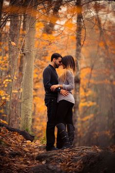 a man and woman are standing in the woods with autumn leaves on the ground behind them