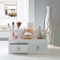 a white drawer with makeup and other items in it next to a vase on a table