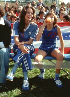 two women sitting on a bench with an american flag in the background and people watching