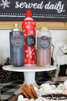 three bottles of hot chocolate on a cake stand with marshmallows and pretzels