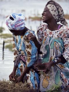 two women standing next to each other in front of water and grass on the ground