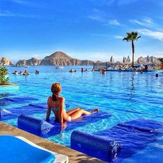 a woman sitting on the edge of a swimming pool in front of mountains and palm trees