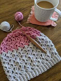 a crocheted hat sitting on top of a wooden table next to a cup of coffee