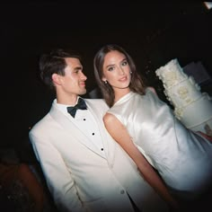 a man and woman standing next to each other in front of a white wedding cake