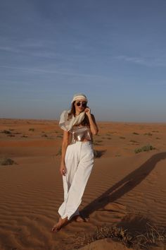 a woman standing in the desert wearing white pants and a cropped top with her hands on her hips