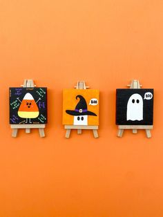 three small eases with halloween paintings on them against an orange wall, one has a ghost and the other has a pumpkin