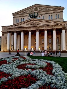 people are sitting on the grass in front of a building with columns and flowers around it