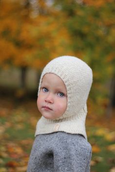 a young boy wearing a white knitted hat and sweater looking at the camera with autumn leaves in the background