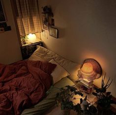 an unmade bed in a dimly lit room with plants on the floor and a lamp next to it