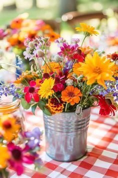 colorful flowers are in a metal bucket on a checkered tablecloth with mason jars