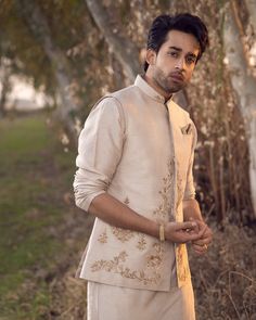 a man standing in front of some trees wearing a white suit and gold bracelets