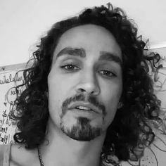 a man with long curly hair wearing a necklace and looking at the camera while standing in front of a wall