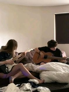 two young people sitting on a bed playing guitar and singing to someone else in the background