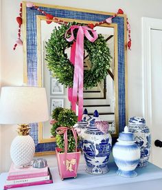 a blue and white table topped with vases next to a wall mounted mirror covered in a pink ribbon