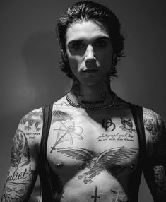 a shirtless man with tattoos on his chest and cross around his neck, standing in front of a white wall