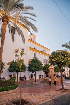 people are walking around in front of a white building with yellow trim and palm trees