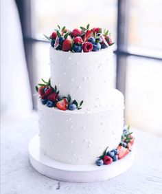 three tiered white cake with berries and blueberries on the top, sitting on a table in front of a window