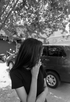 a woman talking on her cell phone while standing in front of a parked car under a tree