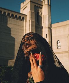 a woman with white nails holding a rose in front of a large building and wearing a veil