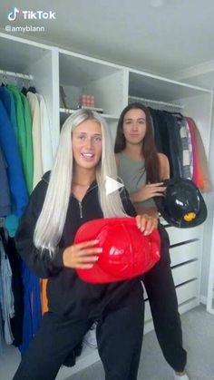 two women standing in front of a closet with hats on their heads and one holding a red object