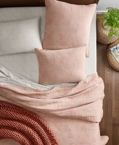 a bed covered in pink linens and pillows next to a basket with green plants