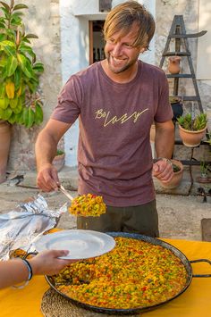 a man is serving food out of a large pan on a table with yellow cloth