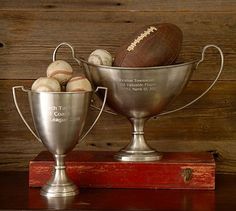 two silver cups with baseballs and a football in it on top of a wooden table