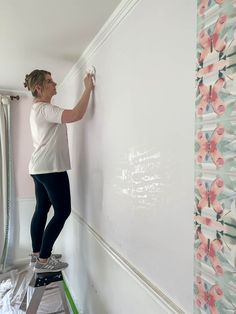 a woman is painting the wall with white paint and she is standing on a ladder