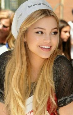 a woman with long blonde hair wearing a white hat and smiling at the camera while standing in front of a group of people