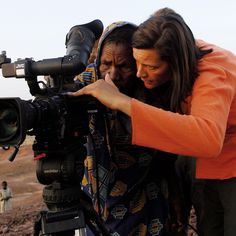 a woman holding a camera up to her face while standing next to a man in an orange shirt