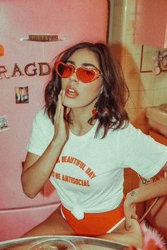 a woman wearing red sunglasses sitting in front of a pink refrigerator with her hand on her face