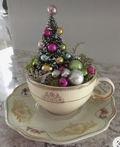 a small christmas tree in a teacup on a saucer, with ornaments around it