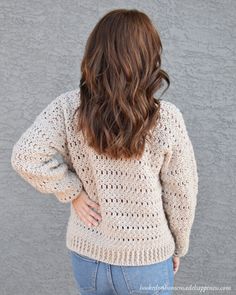 a woman standing in front of a wall wearing a crocheted sweater and jeans