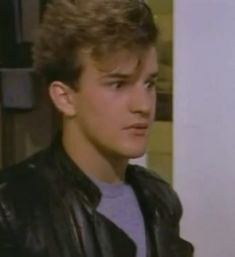 the young man is wearing a leather jacket and looking off into the distance while standing in front of a wall