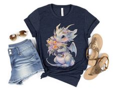 Our dragon with flower shirt is a great gift for those who love baby dragons and flowers. The comfortable fit and cool design of this dragon shirt means that it feels just as good to wear as it is to look at. If you've been looking for a dragon t-shirt for yourself or looking for a dragon lover gift for your best friend, you should get this super cute dragon shirt today. Bella + Canvas Brand Shirt ▶Unisex Adult Sizing - this size runs slightly large. If you want a more fitted look please size down. ▶See Our Size Chart For Proper Sizing ▶Rolled sleeves are for styling purposes only ▶Props used in photos are not included with the purchase. ✈ PROCESSING & SHIPPING ✈ Processing Time: 1-3 business days Standard Shipping: 2-5 business days after processing time ❤ Returns & Exchanges ❤ We know yo Summer T-shirt With Cartoon Print For Gift, Summer Cartoon Print T-shirt For Gift, Summer Cartoon Print T-shirt As A Gift, Casual Cartoon Print T-shirt For Gifting, Casual Cartoon Print T-shirt For Gift, Casual T-shirt With Cartoon Print For Gift, Casual Tops With Character Print For Gifts, Casual T-shirt With Character Print As Gift, Casual T-shirt With Character Print For Gift