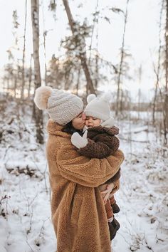 a woman in a brown coat holding a baby while standing in the snow with trees behind her