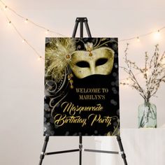 a black and gold masquerade birthday party sign on an easel with lights in the background