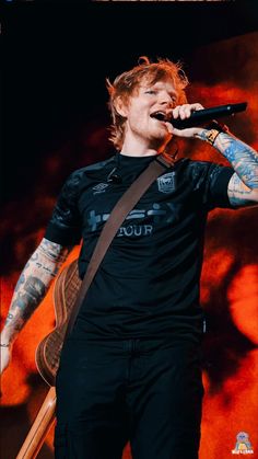 a man with tattoos on his arm holding a guitar and singing into a microphone in front of an orange background