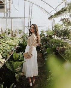 a woman in a white dress standing in a greenhouse with lots of plants on the shelves