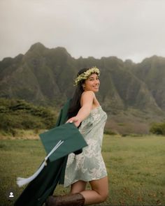 a woman in a green dress is holding her graduation cap and gown with mountains in the background