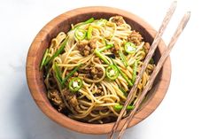 a wooden bowl filled with noodles and meat next to chopsticks on a white surface